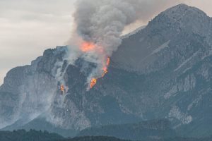 Wildfire with orange flames and thick smoke plume rising from a rocky mountainside in Peña Montañesa, Huesca, Spain, with forested hills in the foreground