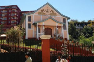 Frontal view of the peach-and-white High Constitutional Court building in Ambohidahy, Antananarivo, featuring a gold scales-of-justice bas-relief on its gabled façade, a wrought-iron fence in the foreground, and pedestrians walking along the street under a clear blue sky.