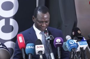 Taiwo Oyedele wearing a blue suit and printed tie, looking down while speaking into a cluster of press microphones.