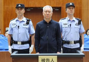 Former China Evergrande Group founder Xu Jiayin standing at the defendant's podium in a Chinese courtroom between two police officers during his sentencing.