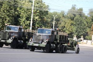 Two green military utility trucks carrying soldiers in camouflage gear and towing towed artillery cannons drive across an asphalt street during a public parade in Moldova.