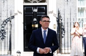 UK Prime Minister Andy Burnham wearing glasses and a dark suit stands outside the black entrance door of 10 Downing Street.