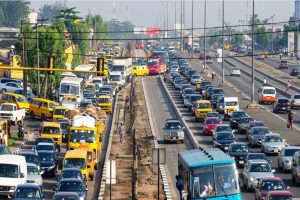 A high-angle view of a multi-lane highway in Lagos crowded with cars, yellow commercial minibuses, and blue and red BRT buses in heavy traffic beside a concrete median walkway.