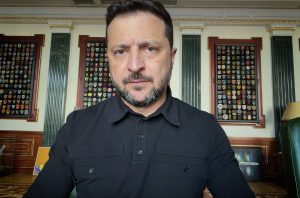 Ukrainian President Volodymyr Zelenskyy seated in his office wearing a black long-sleeve shirt, looking directly into the camera with framed displays of Ukrainian military unit patches on the wall behind him.